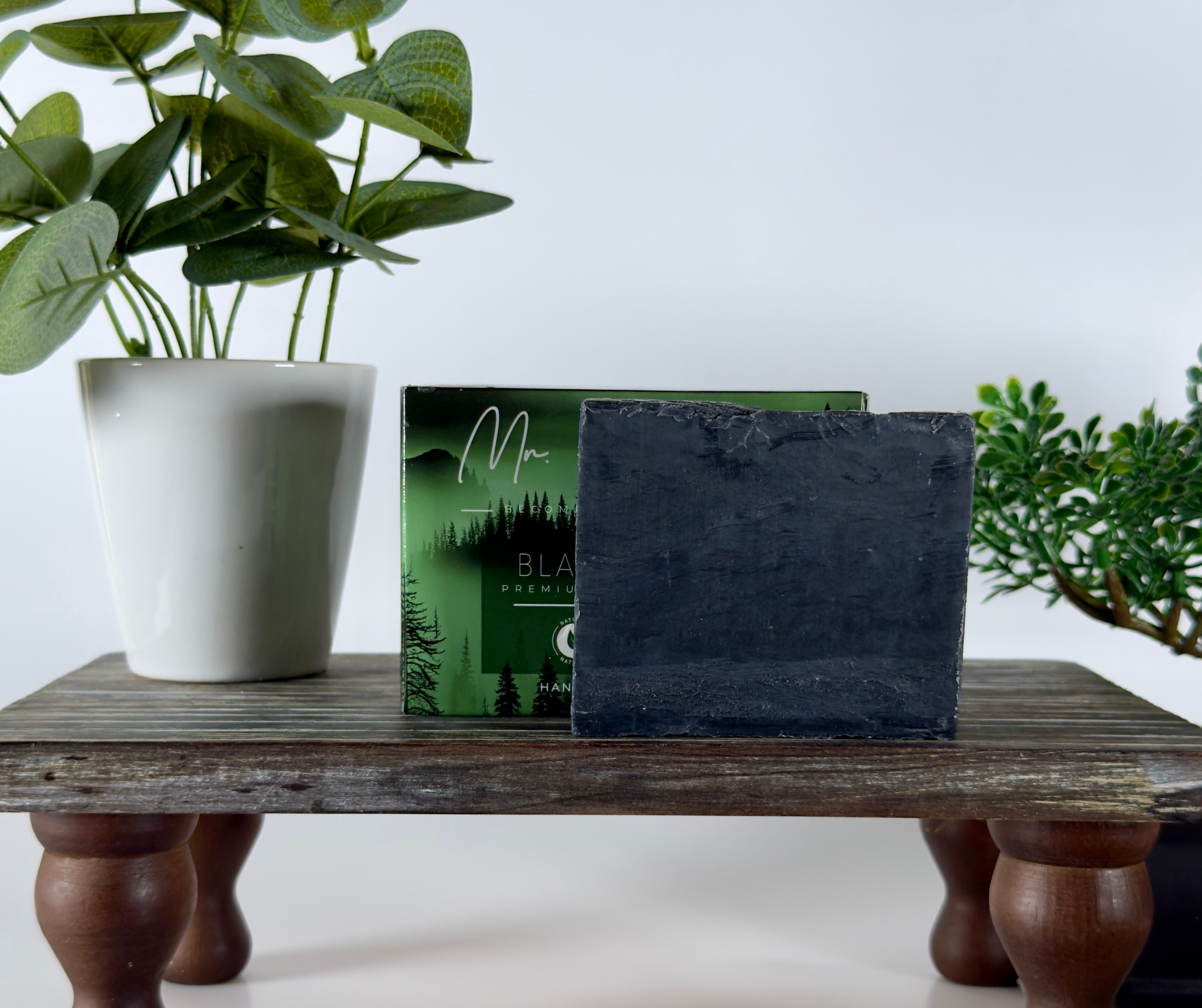 Black soap bar and packaging on a wooden surface with plants in the background